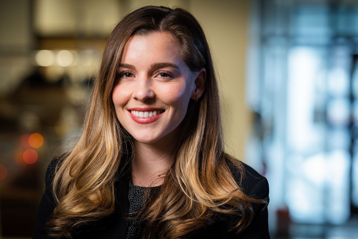 Professional portrait with soft lighting shows a warm smile and long blonde ombre hair against a blurred office background.