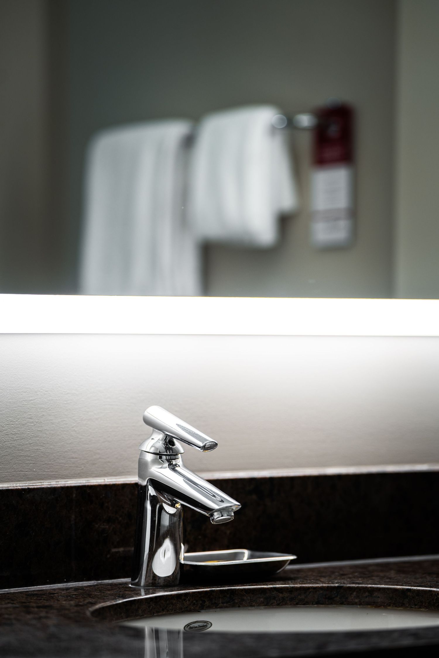 Modern chrome bathroom faucet on black granite countertop with white towels hanging in background.