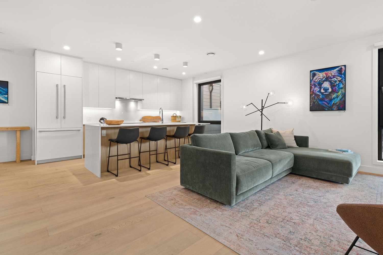 Modern open concept living room with green sectional sofa, hardwood floors and kitchen with breakfast bar in background.
