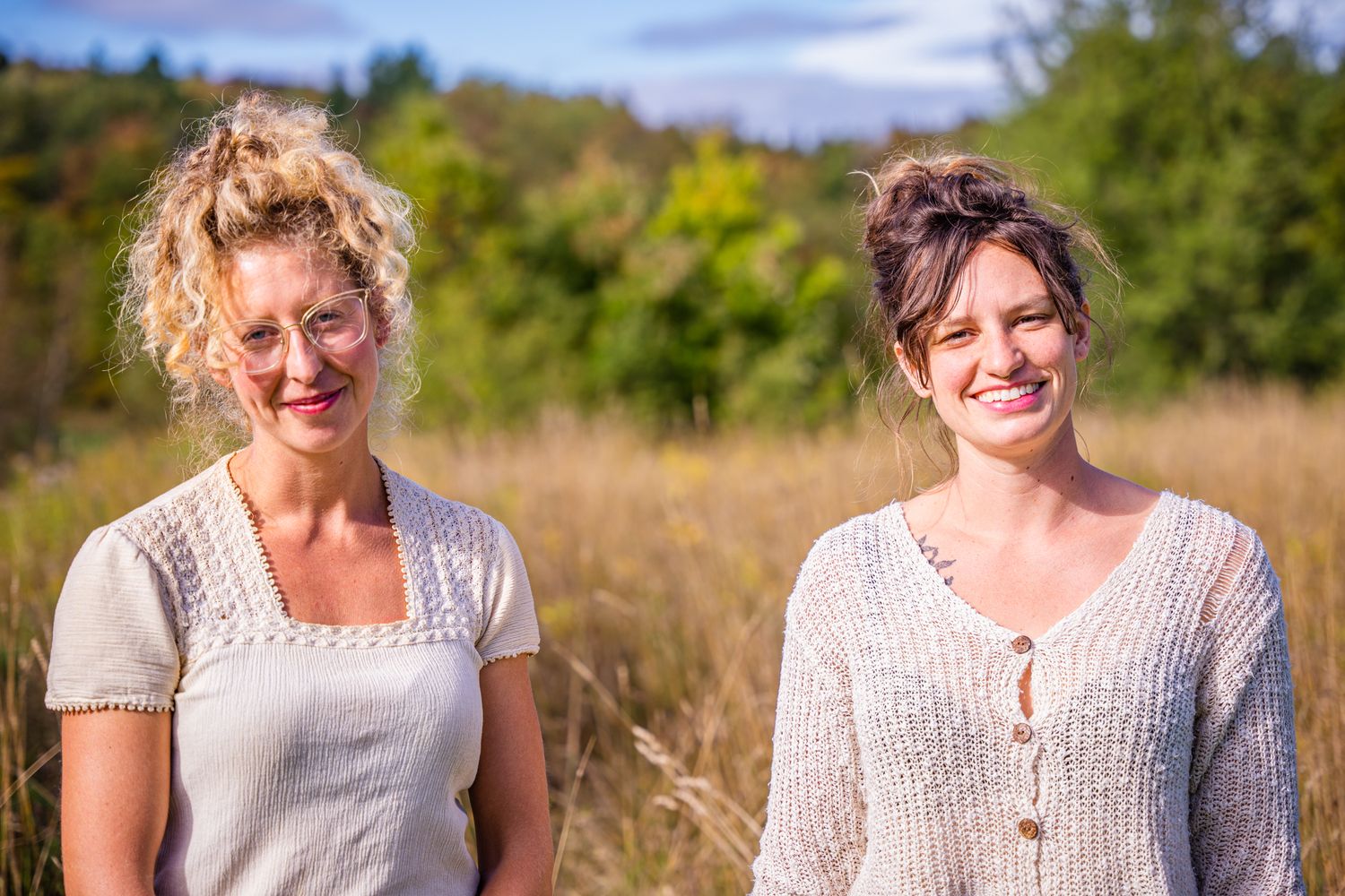 Women in white tops and denim sitting together in a grassy meadow on a bright summer day.