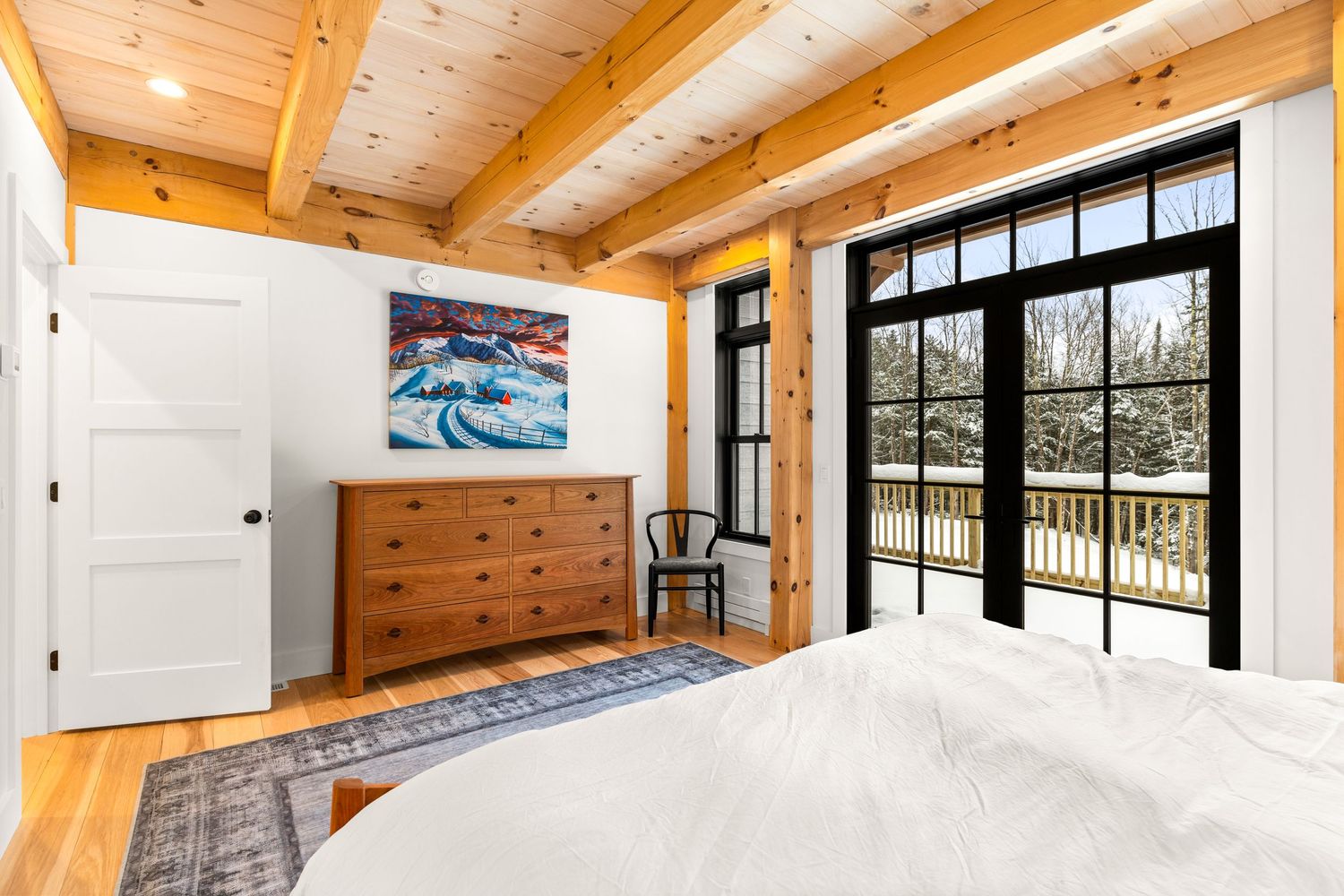 Cozy bedroom with exposed wooden beams, large black-framed windows, and a watercolor print on the wall.