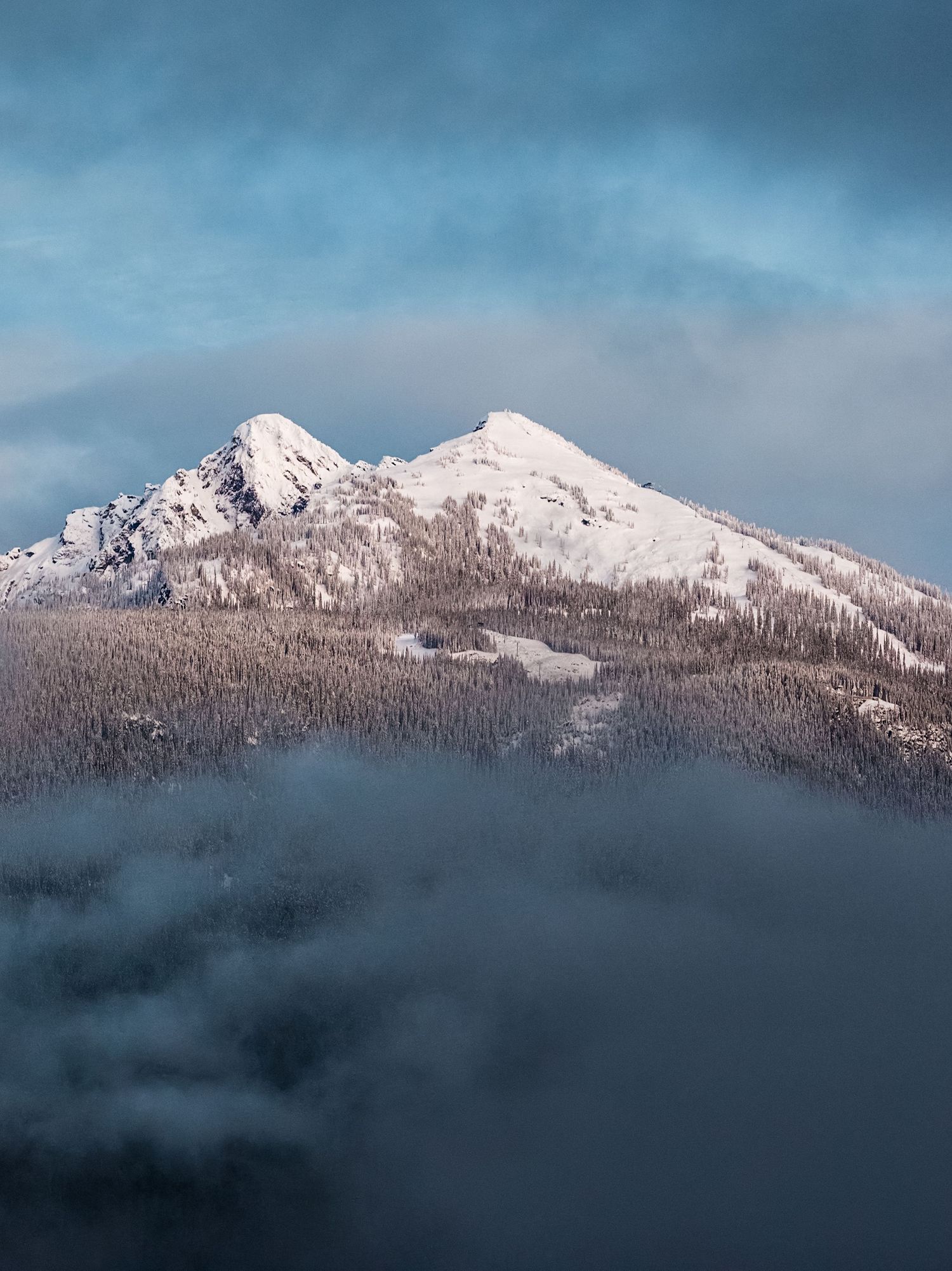 Snow-capped mountain peaks emerge through misty clouds against a moody blue winter sky.