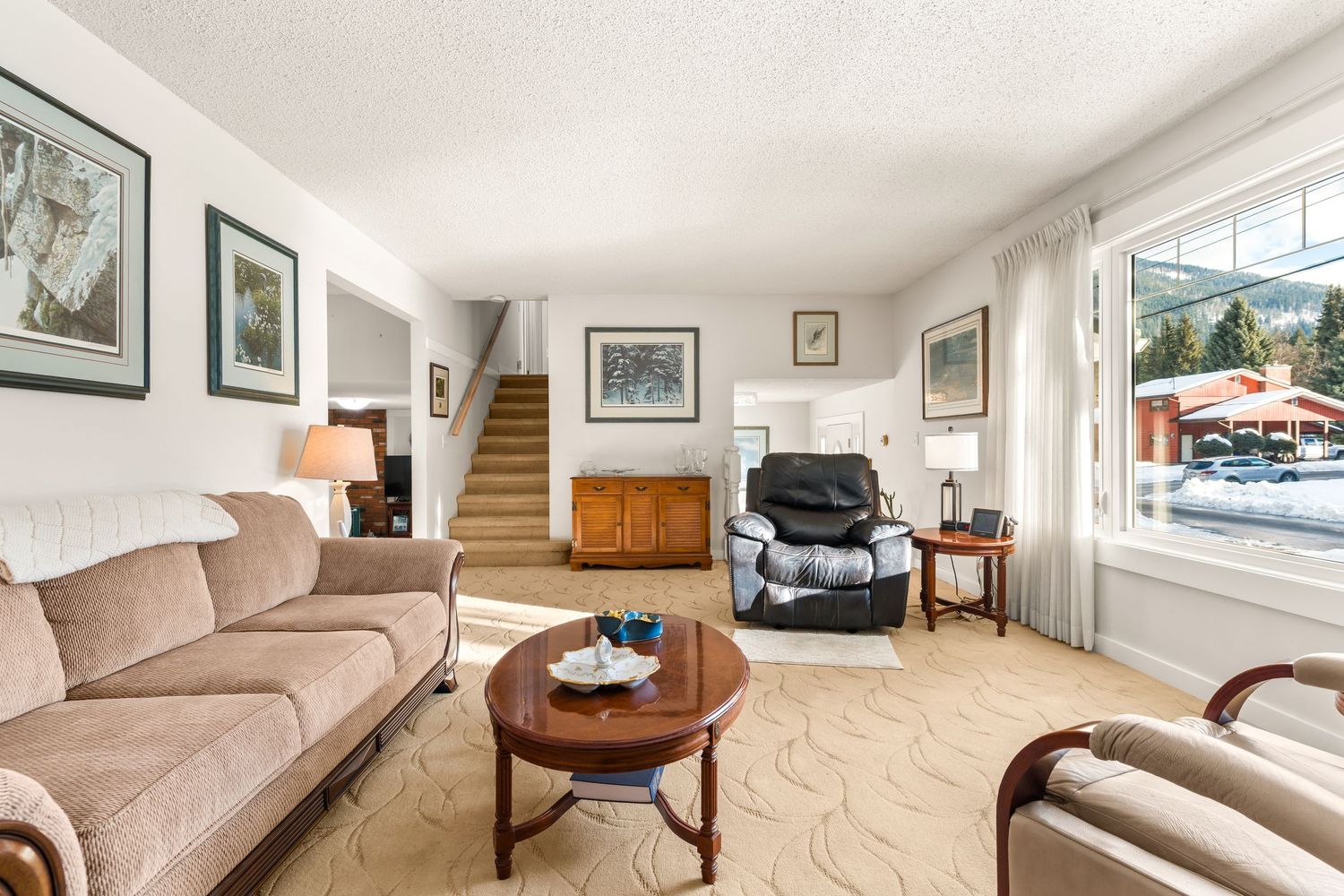 Bright living room with beige sofa, round coffee tables, recliner chair and carpeted staircase leading upstairs.