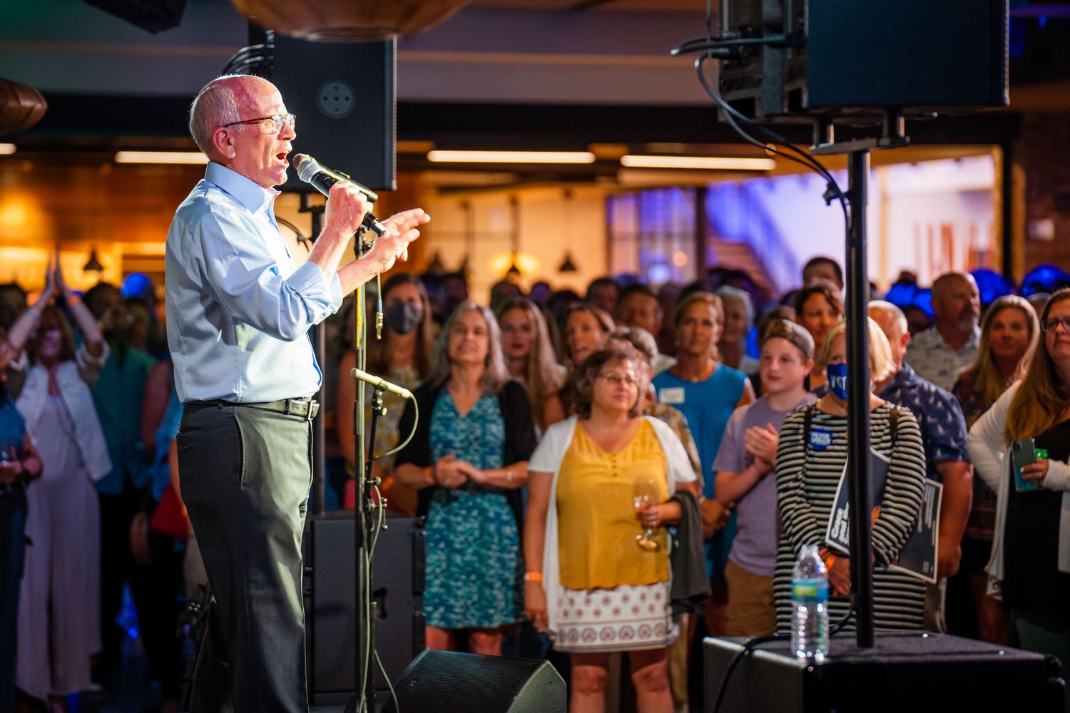 A speaker presents to an engaged audience at a business conference in a warmly lit indoor venue.