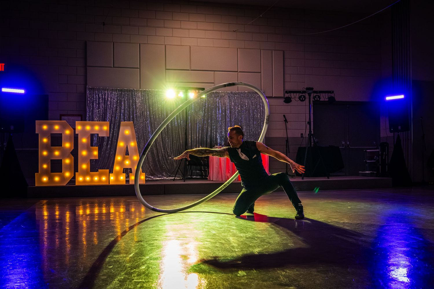 Performer in silhouette dances with a hula hoop on dark stage with illuminated BEAT sign and colorful spotlights.