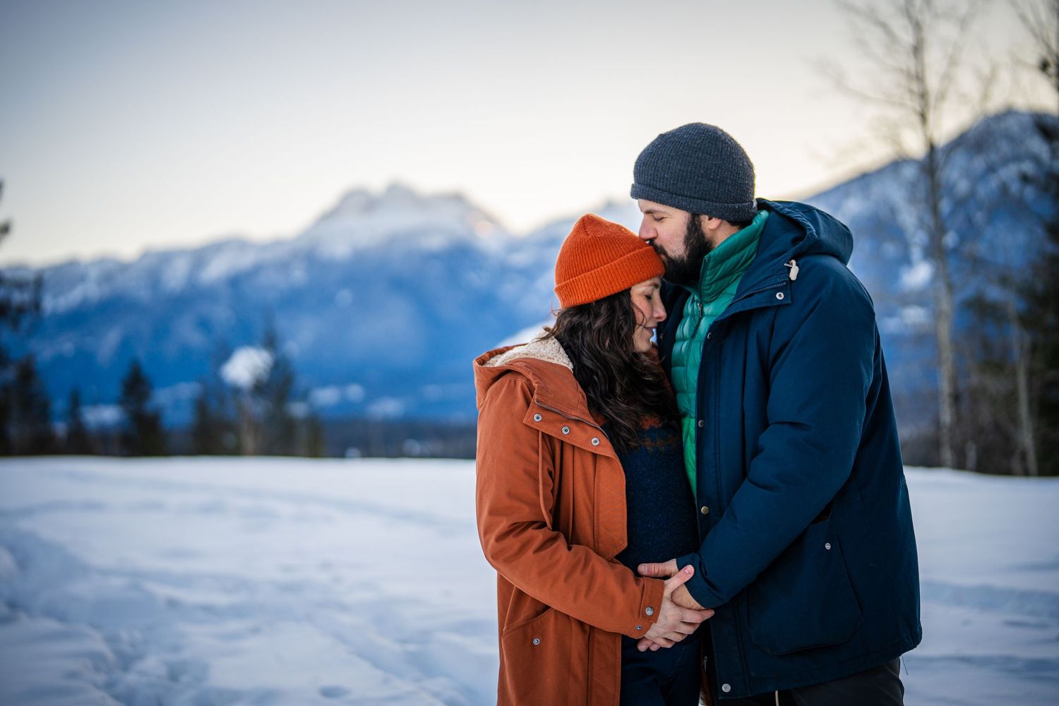 A couple embraces in a snowy winter landscape with snow-capped mountains in the background.