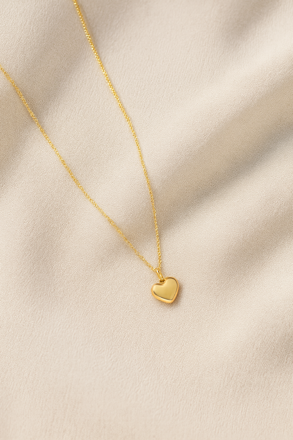 Collar dorado con colgante de corazón sobre fondo crema. Fotografía de producto minimalista para joyería online.