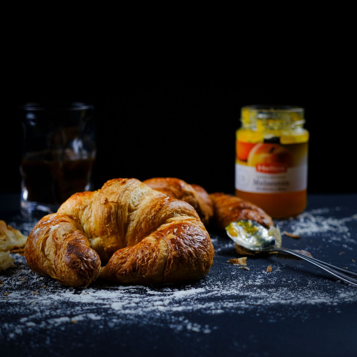 Fotografía gastronómica de croissant artesanal con mermelada y bebida, imagen publicitaria para cafetería.