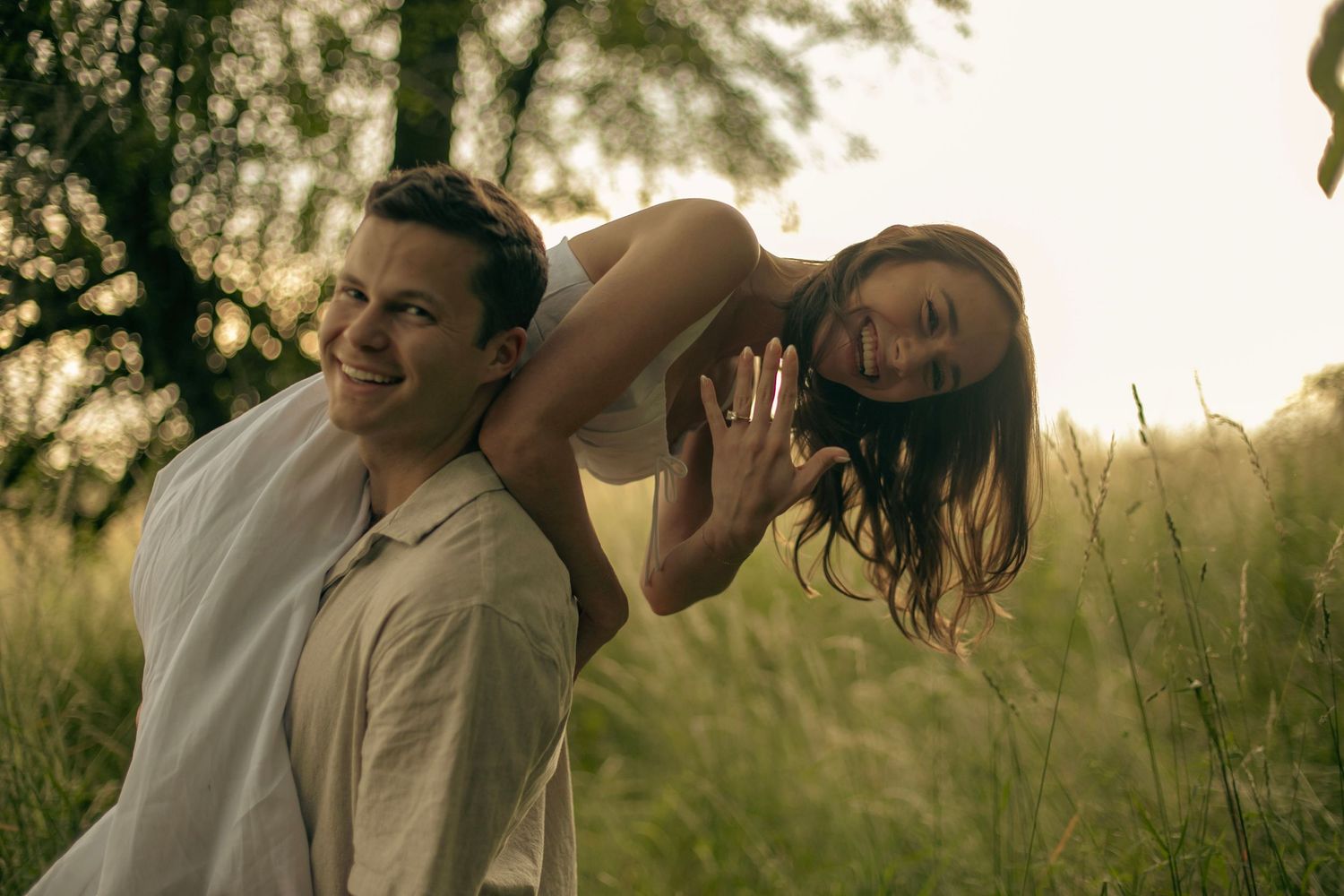 A man holds his fiance over his shoulder while she excitedly shows off her engagement ring in a sunny field of tall grass