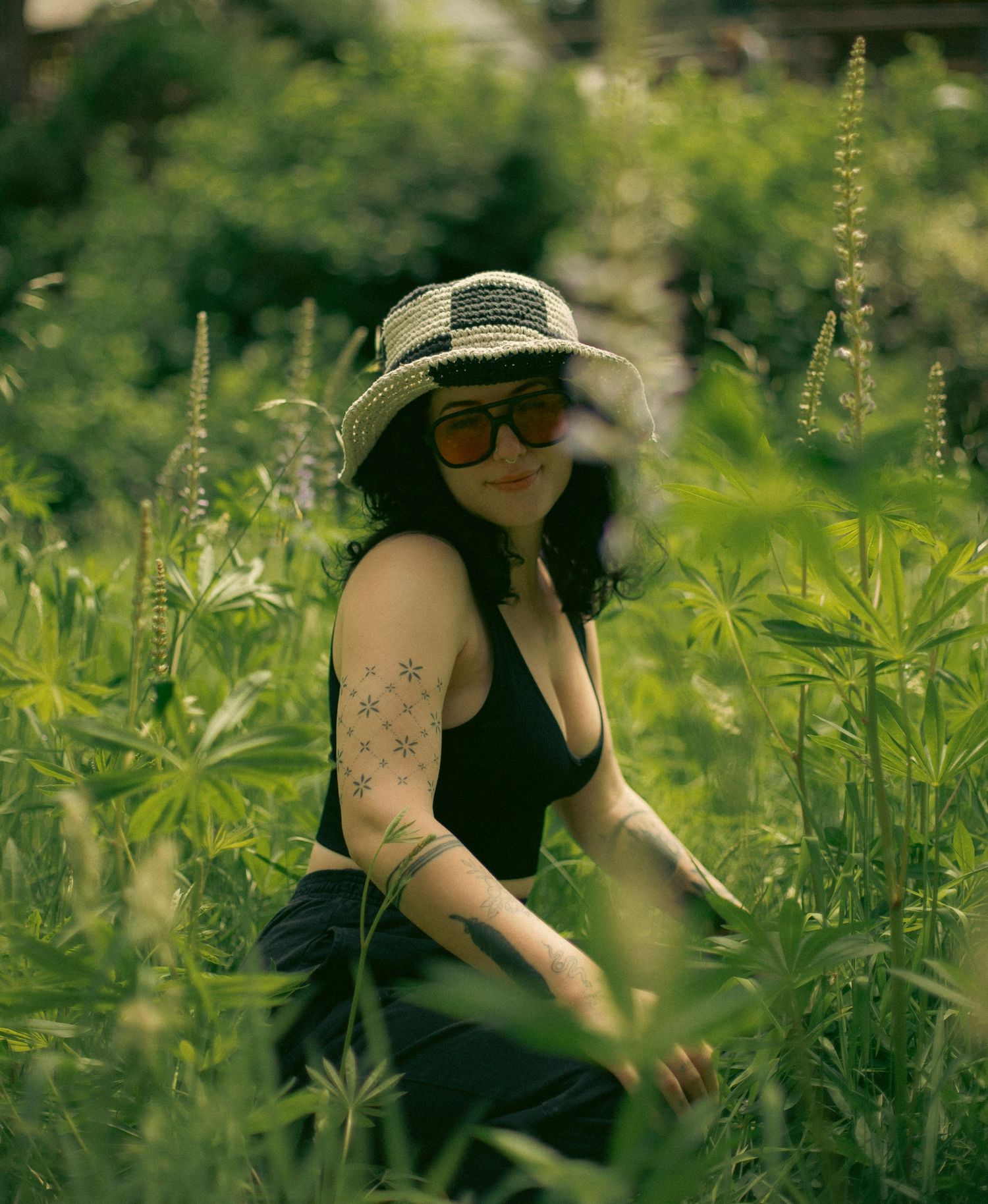 A woman with dark curly hair and a checkered bucket hat sitting in a green field with purple flowers around her.