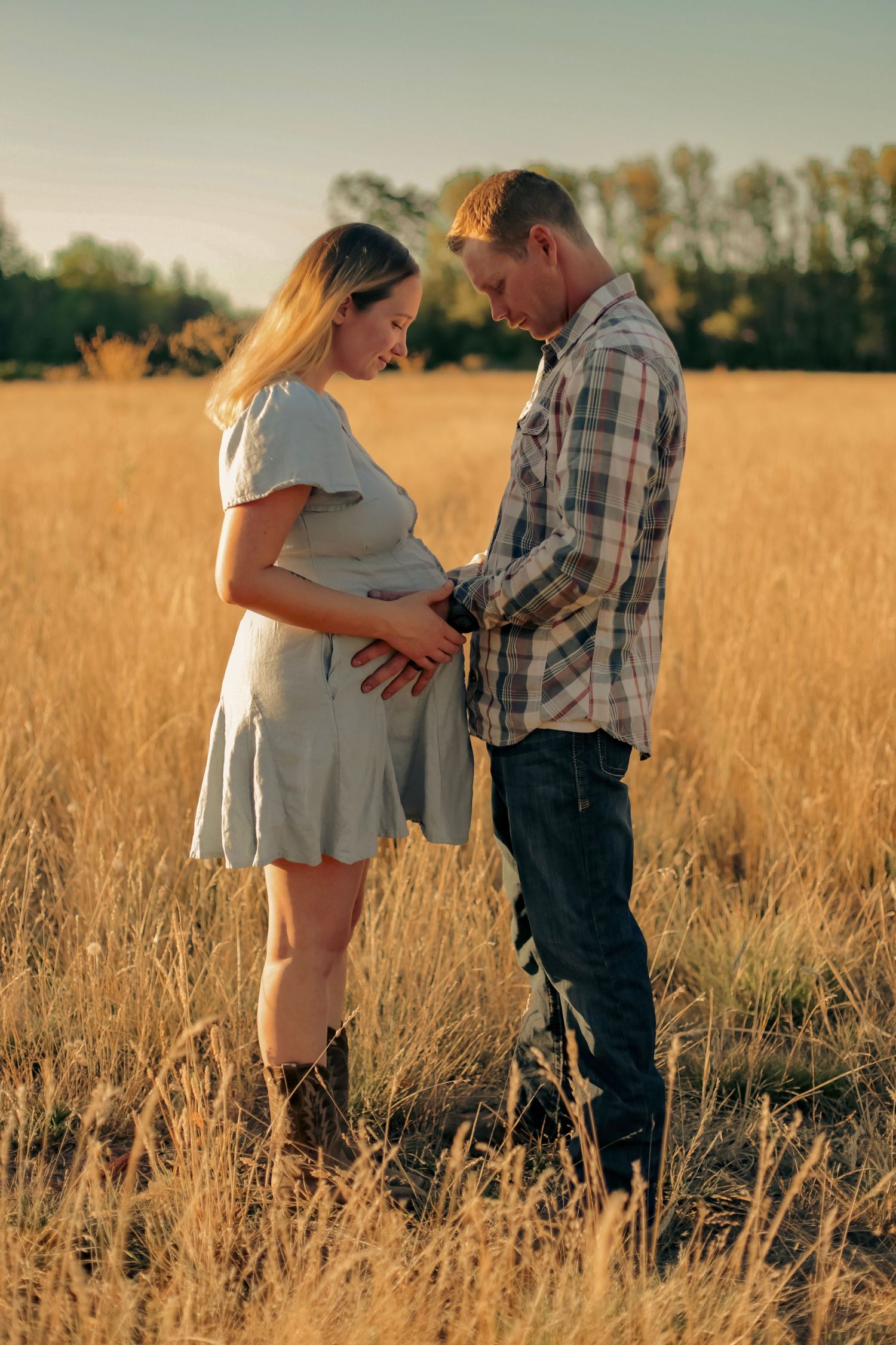 A woman and man facing each other and both holding her pregnant belly while standing in a sunset-lit wheat field.