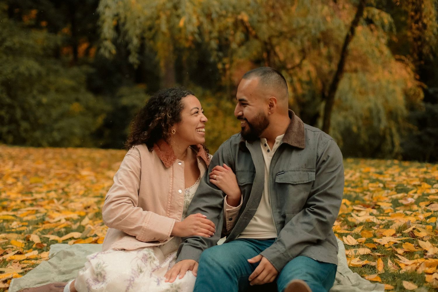 Couple looking at each other lovingly while sitting on a ground covered in yellow leaves with a green and yellow tree in the background.