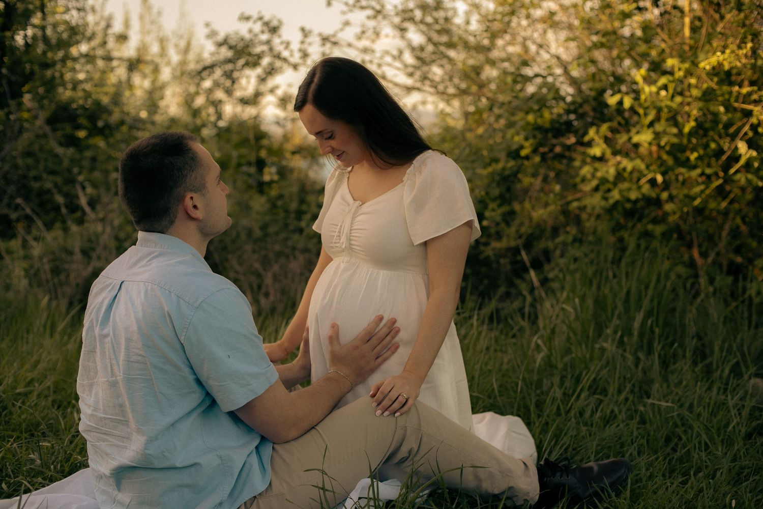 A woman leans against her partner, he holds her belly and looks up lovingly to her while they pose for photos in a cinematic, moody field