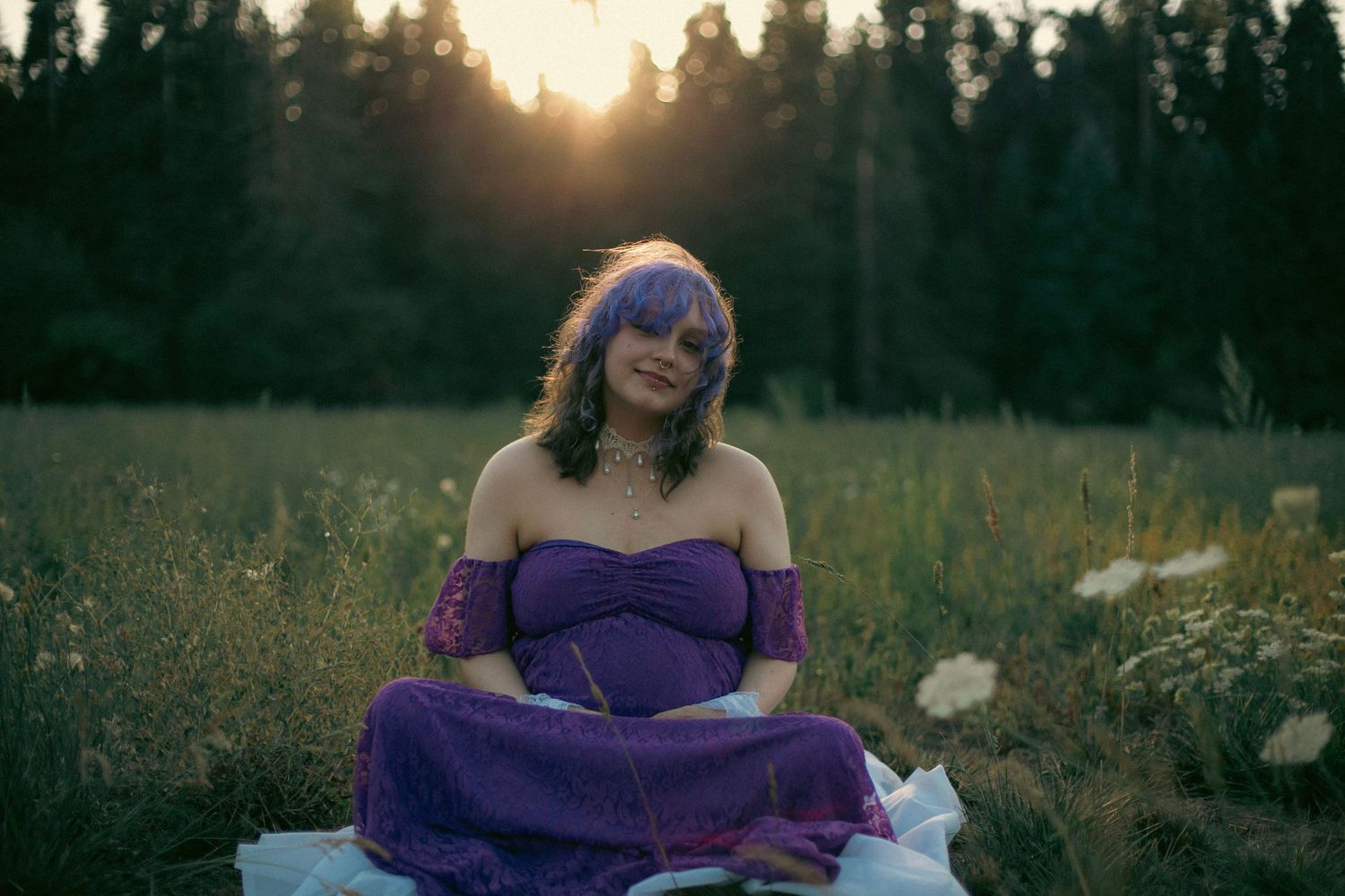 A maternity photo for a woman in a purple dress holding her belly and sitting amongst white flowers in a green grass field.