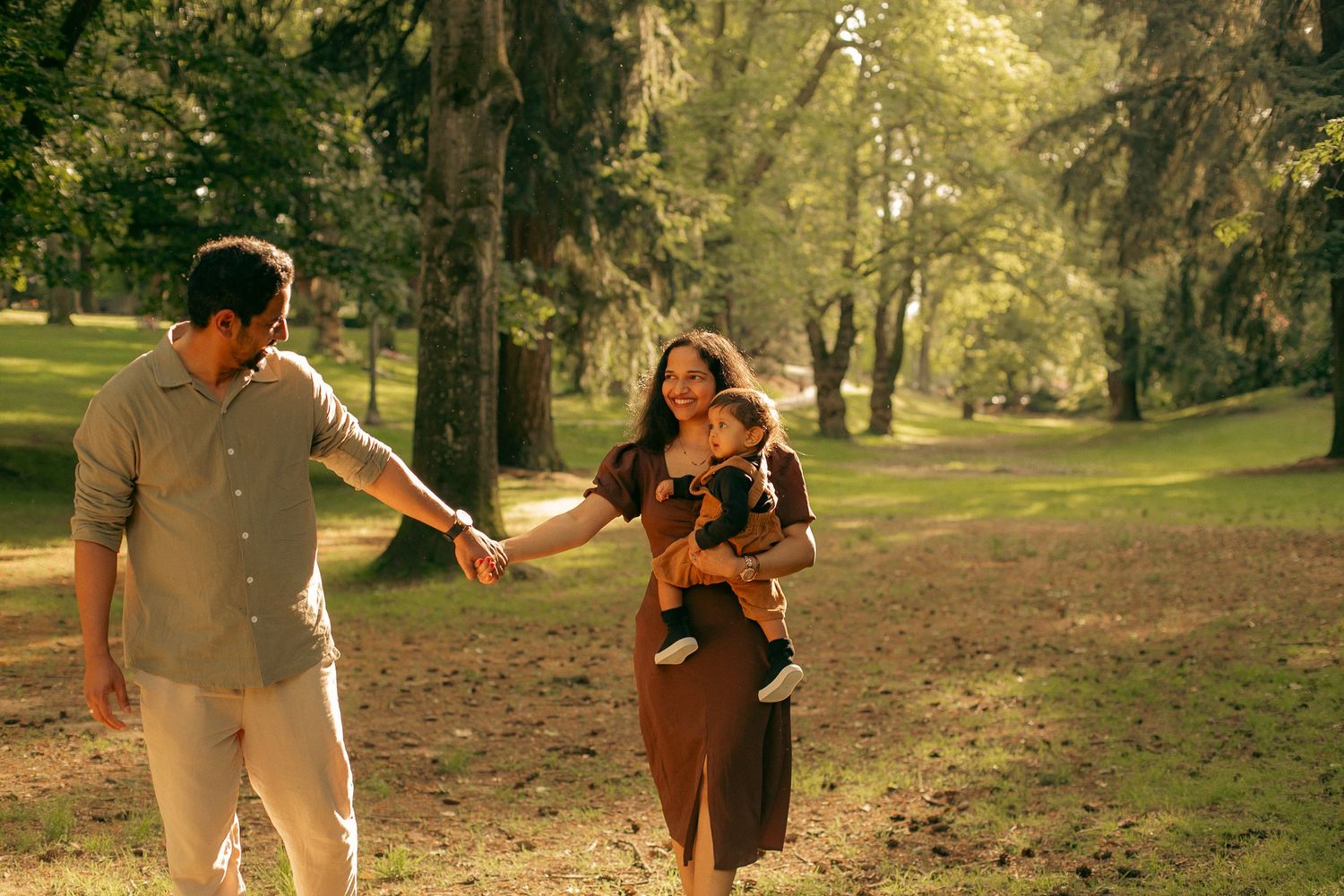 A family of three walk together in a sunny neighborhood park holding hands and smiling