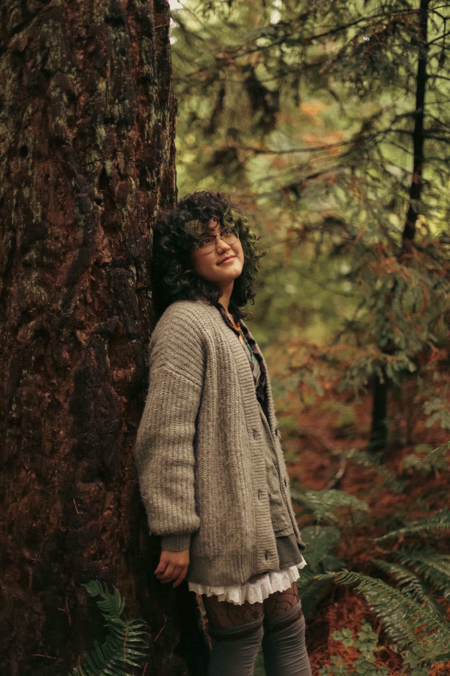 Portrait of a young woman wearing a cozy sweater leaning against a redwood tree in a forest.