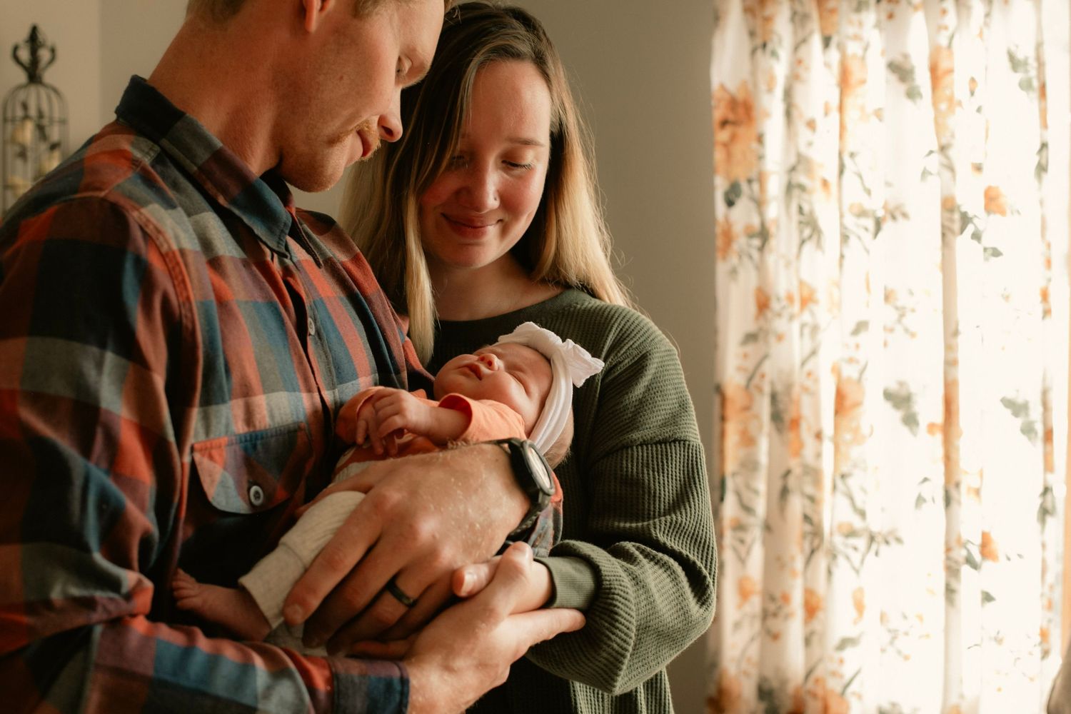 A mother and father holding their newborn baby in a cozy living room with sunlight coming through the window.