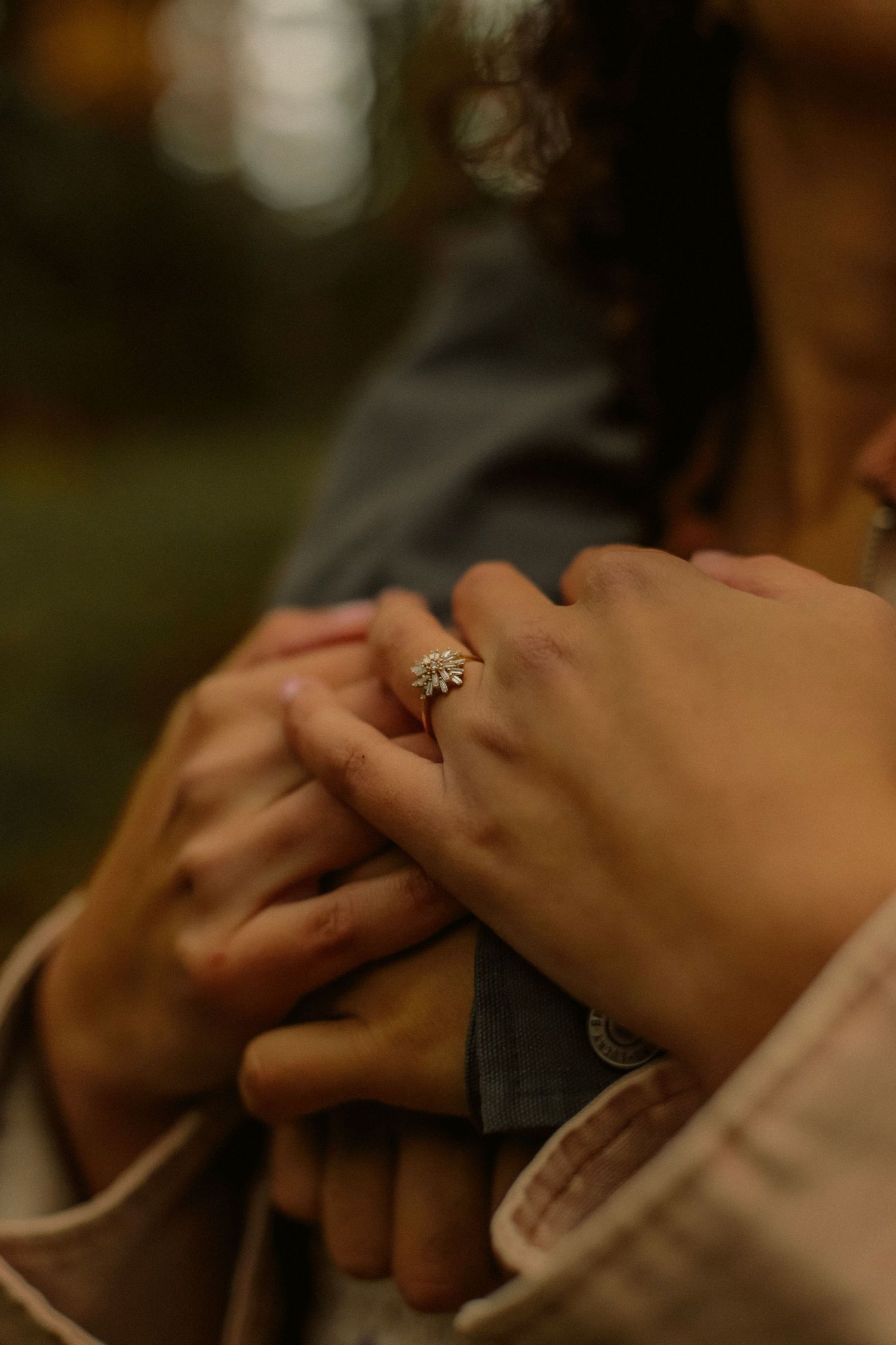 A woman's engagement ring displayed while holding the hands of her fiancé in a moody forest setting.