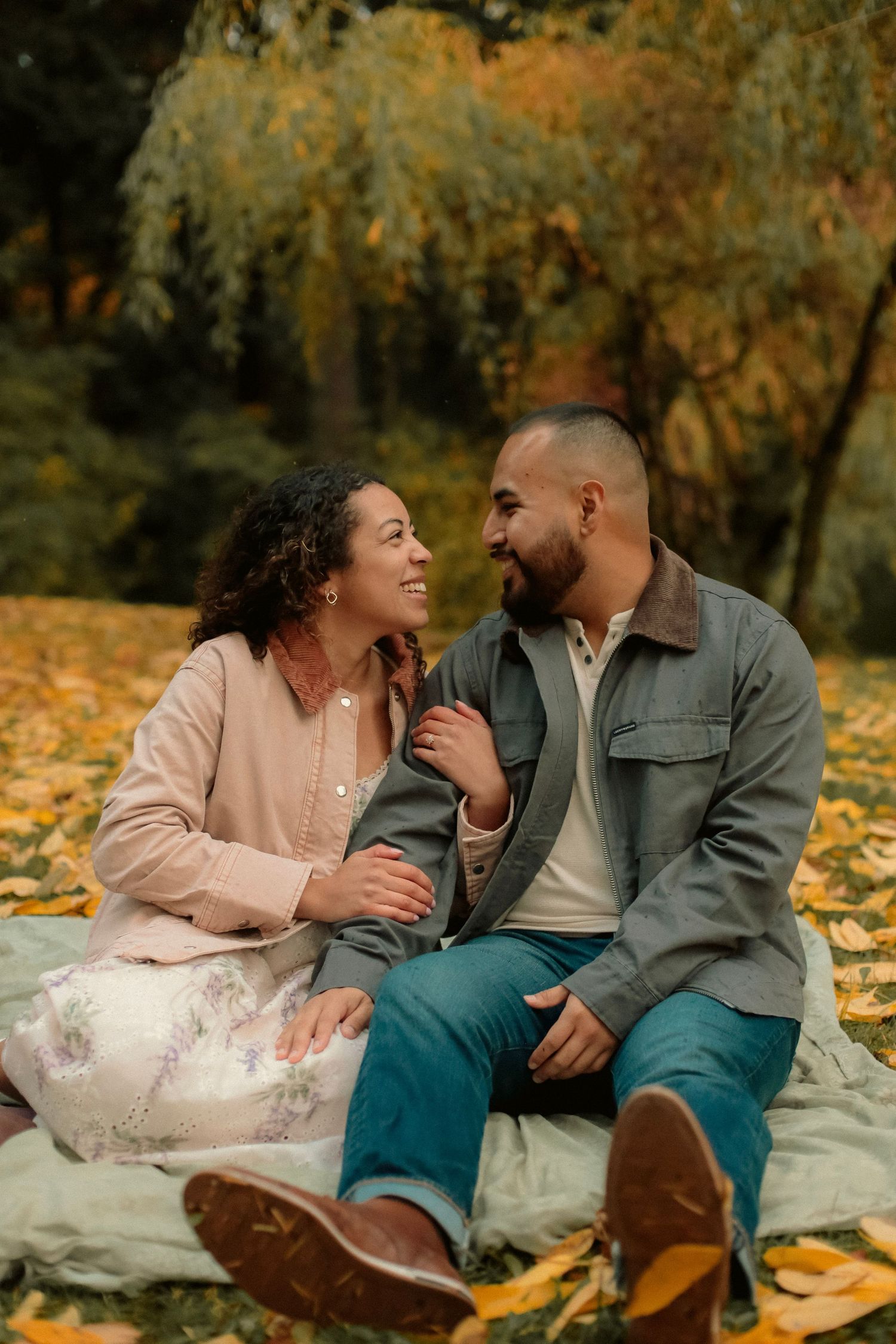 Couple sitting on yellow leaves with a tree in the background. They are looking at each other lovingly and smiling for engagement photos