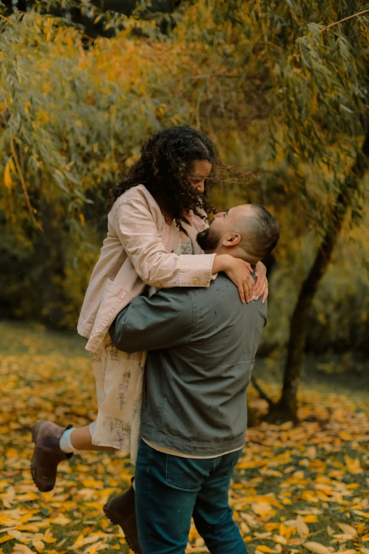 A man picks up his fiancé and swings her around with falling yellow leaves in the background.