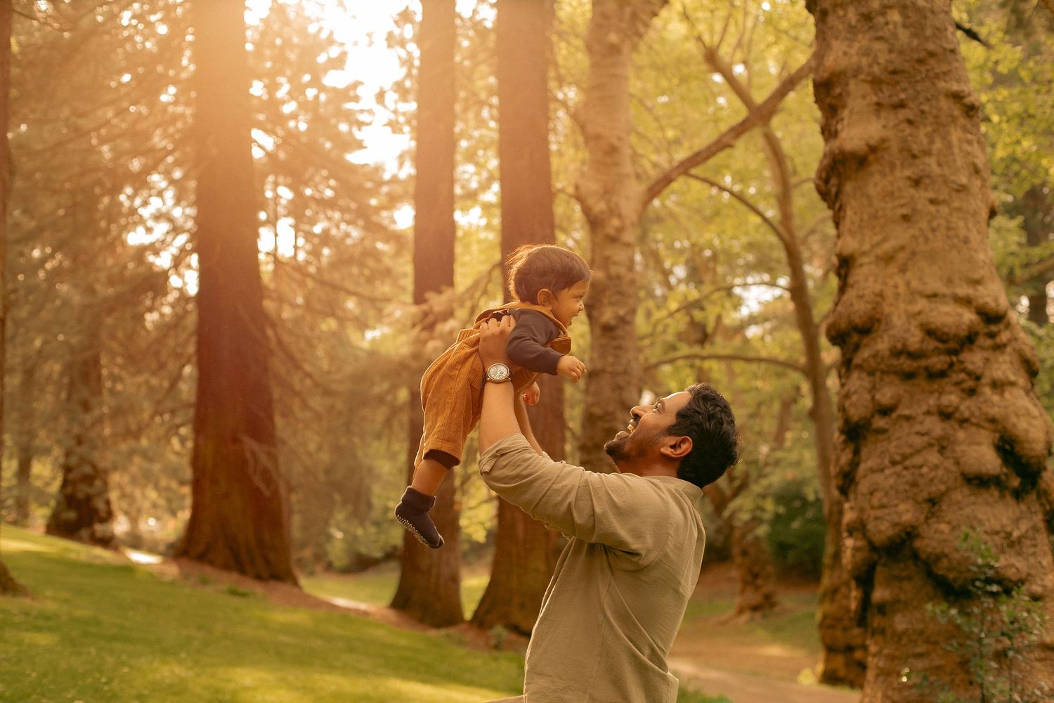 A dad spins his baby in the air and laughs while in a sunny neighborhood park for a family photoshoot