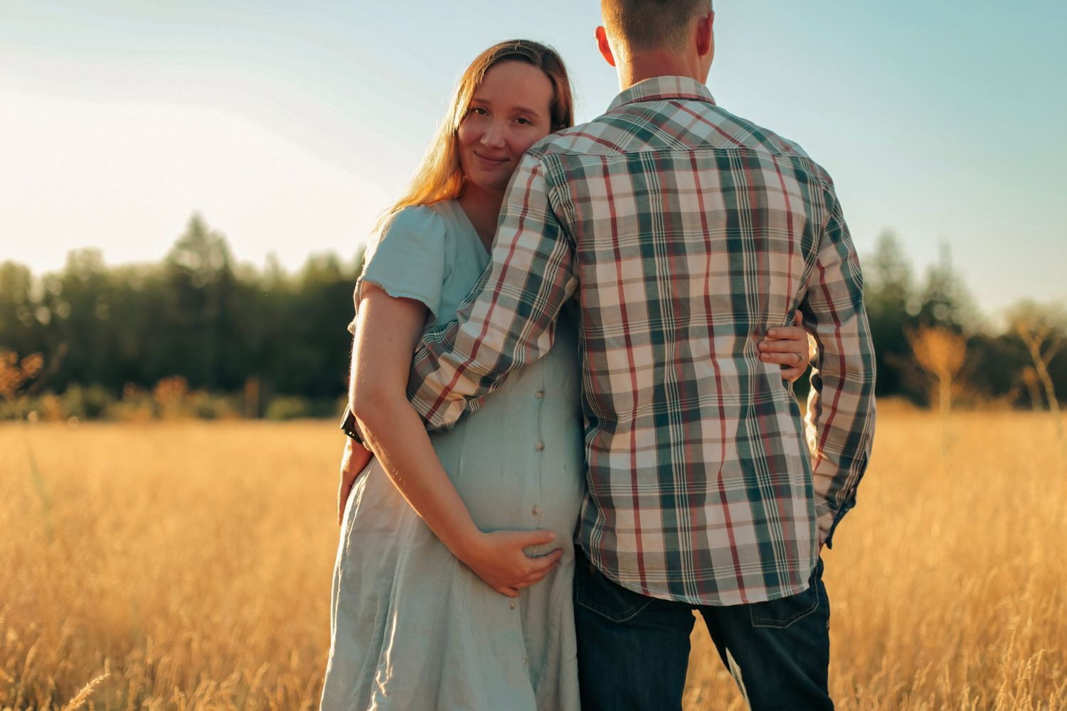 A woman faces the camera holding her belly while her husband holds her and faces the other way