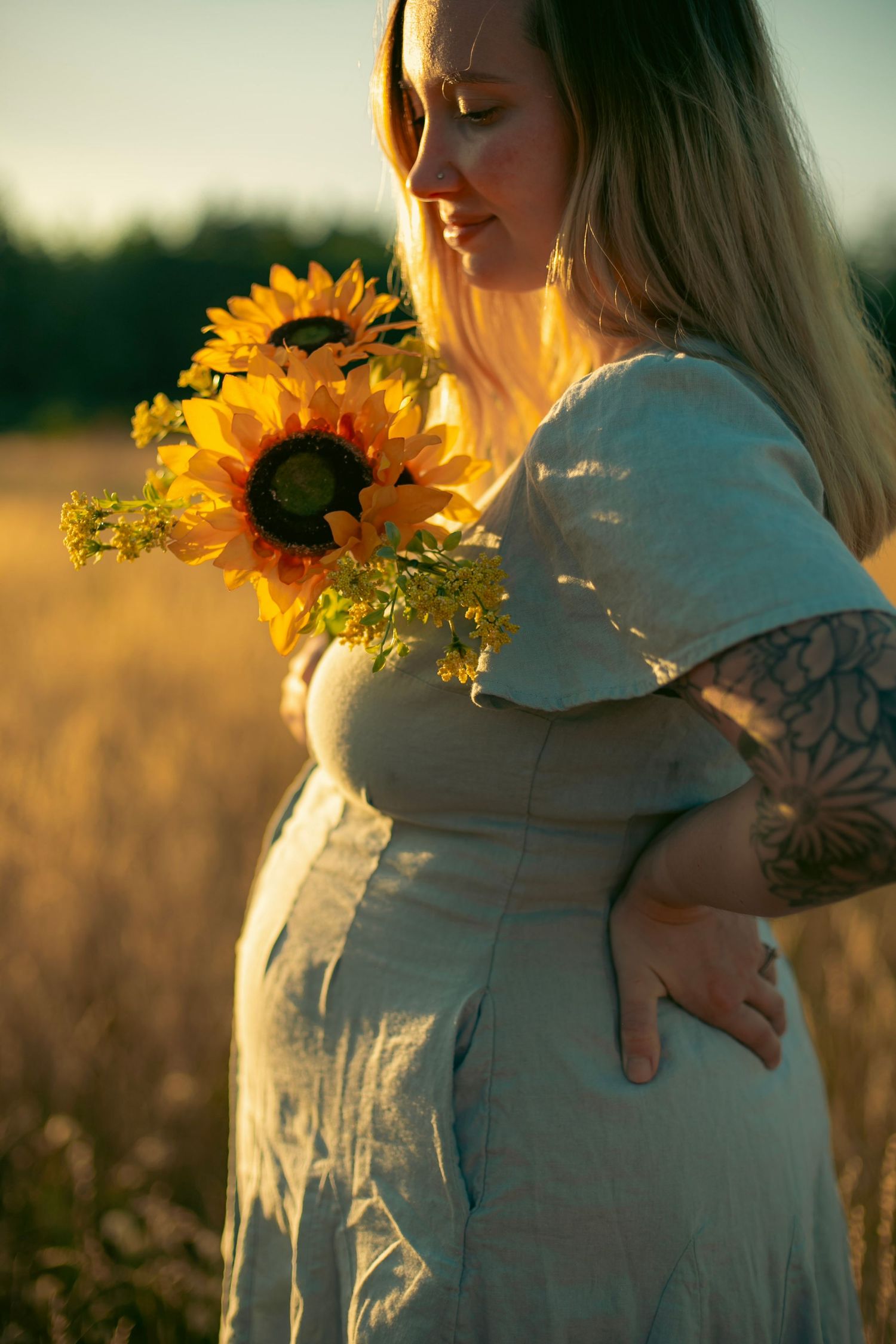 A  pregnant woman with blonde, straight hair in a blue dress holding sunflowers close to her chest while standing in a field.