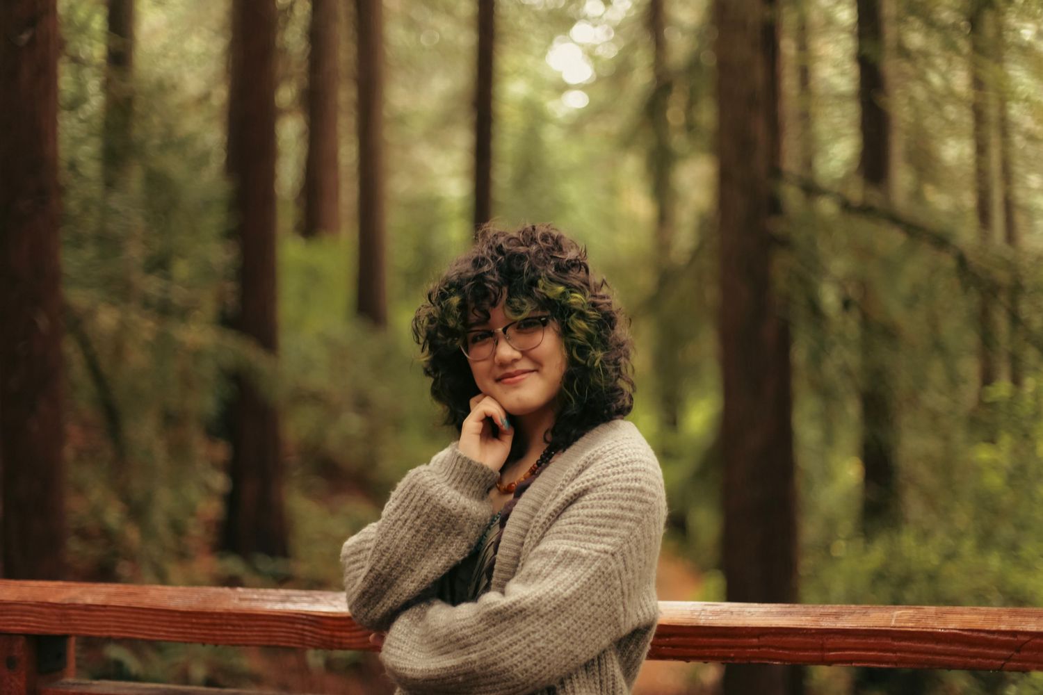 A young woman poses with her hand to her chin with redwood trees in the background for a high school senior graduation photoshoot