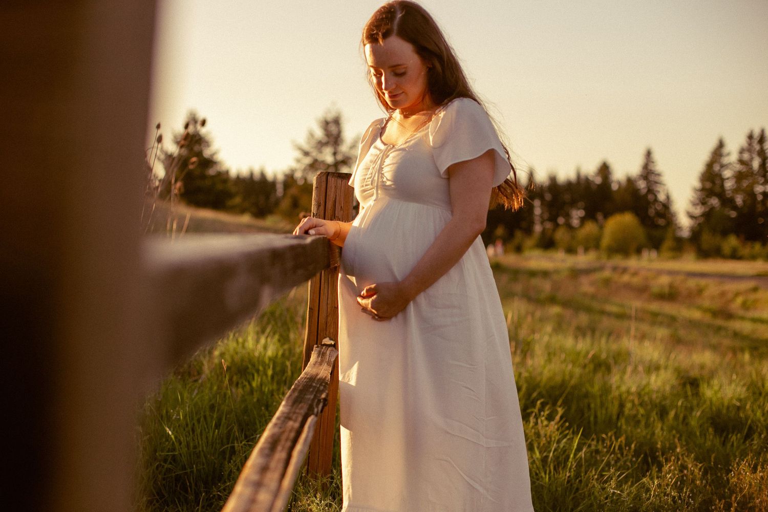 A woman in a white dress stands in a rural field leaning against a wooden fence while holding her pregnant belly for a maternity photoshoot