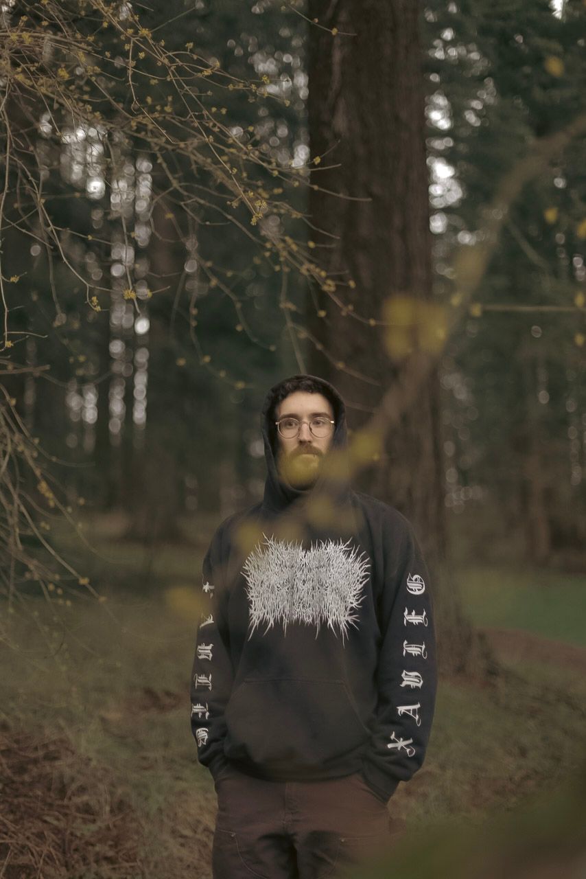 A man with a black sweater standing in a forest with branches in front of him.