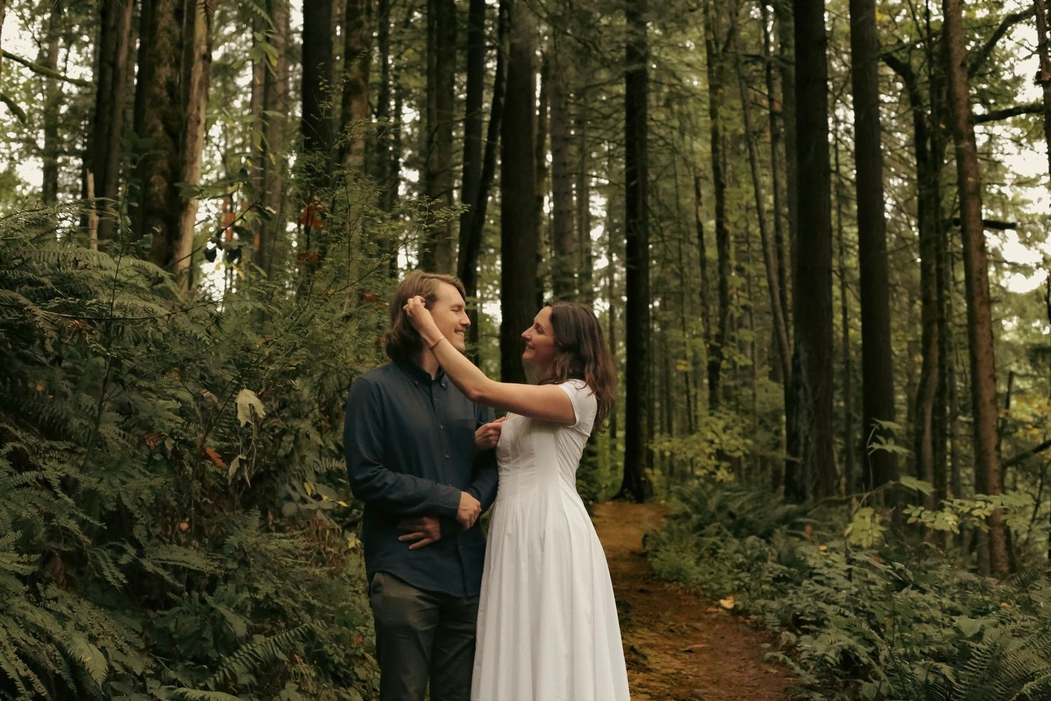 A woman in a white dress brushing the hair back from the face of her partner while looking at each other and smiling in a green, lush forest