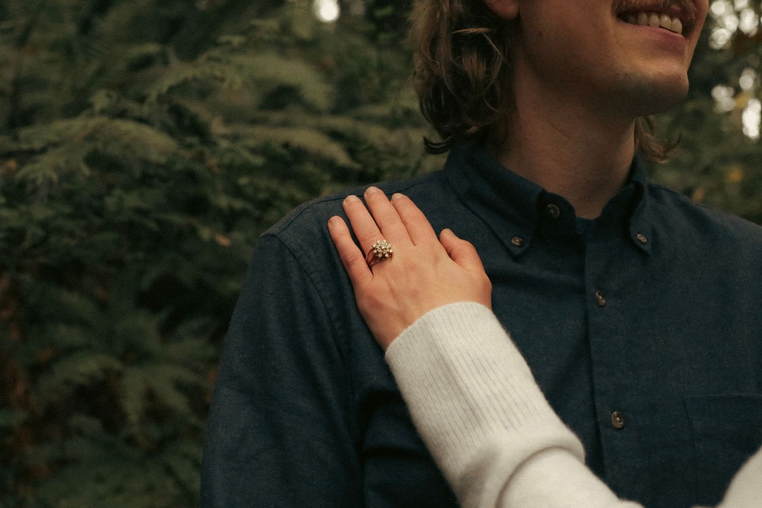 A woman's hand showing her engagement ring on the shoulder of her partner with ferns in the background.