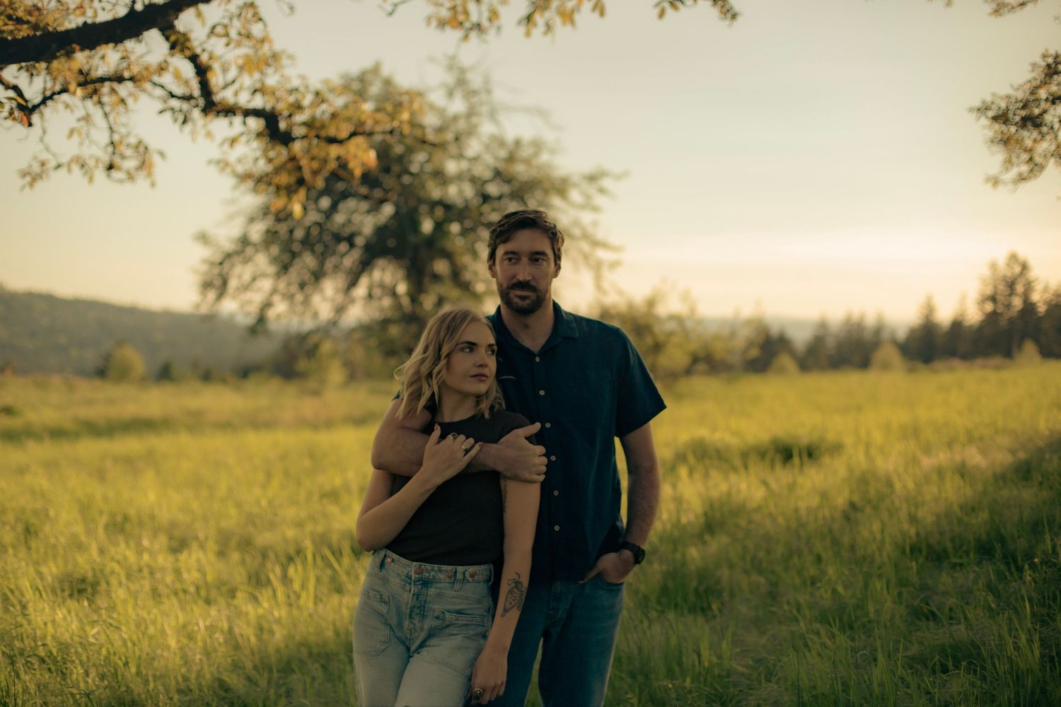 A couple poses in a green grassy field on a summer day, the man holds his arm around the woman while she looks in the other direction