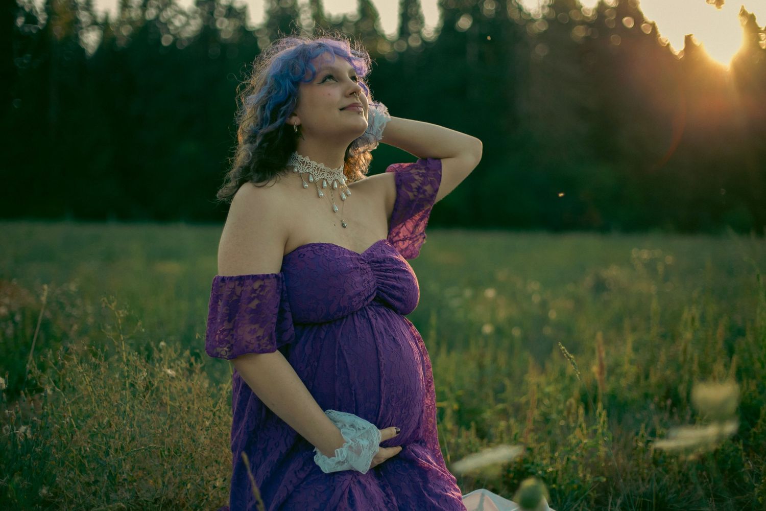 A maternity photo for a woman in a purple dress holding her belly and sitting amongst white flowers in a green grass field.