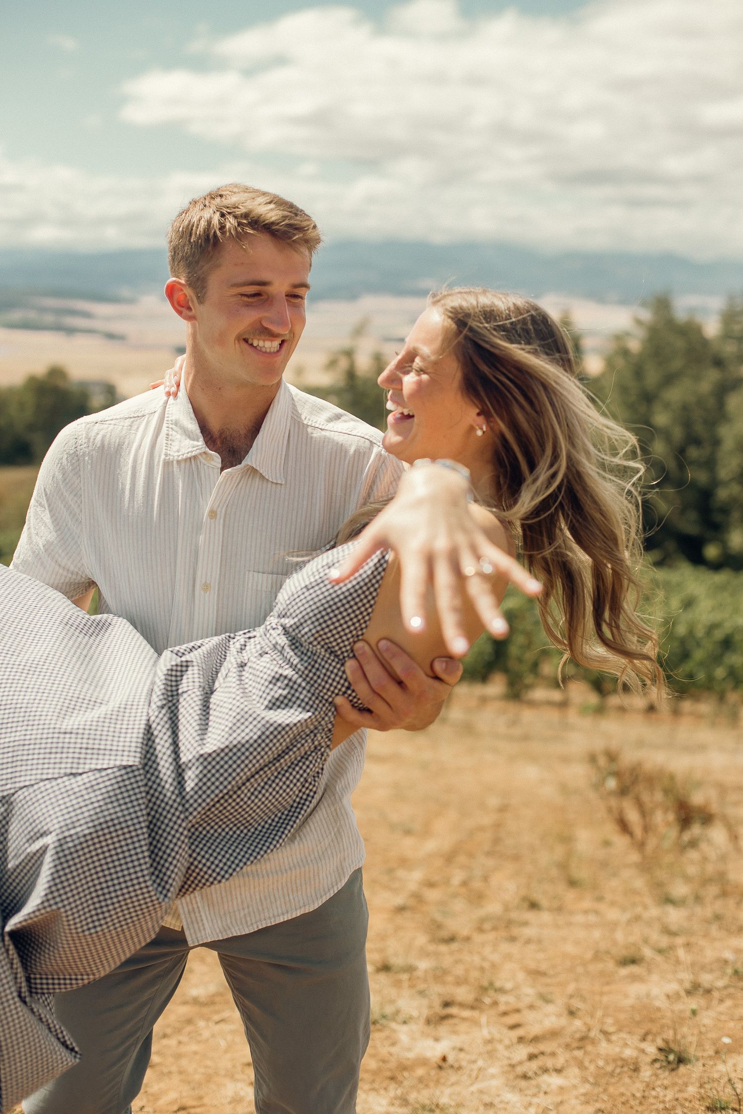 A man picking up his fiancé and twirling her around in a bright, warm vineyard to show off their engagement.