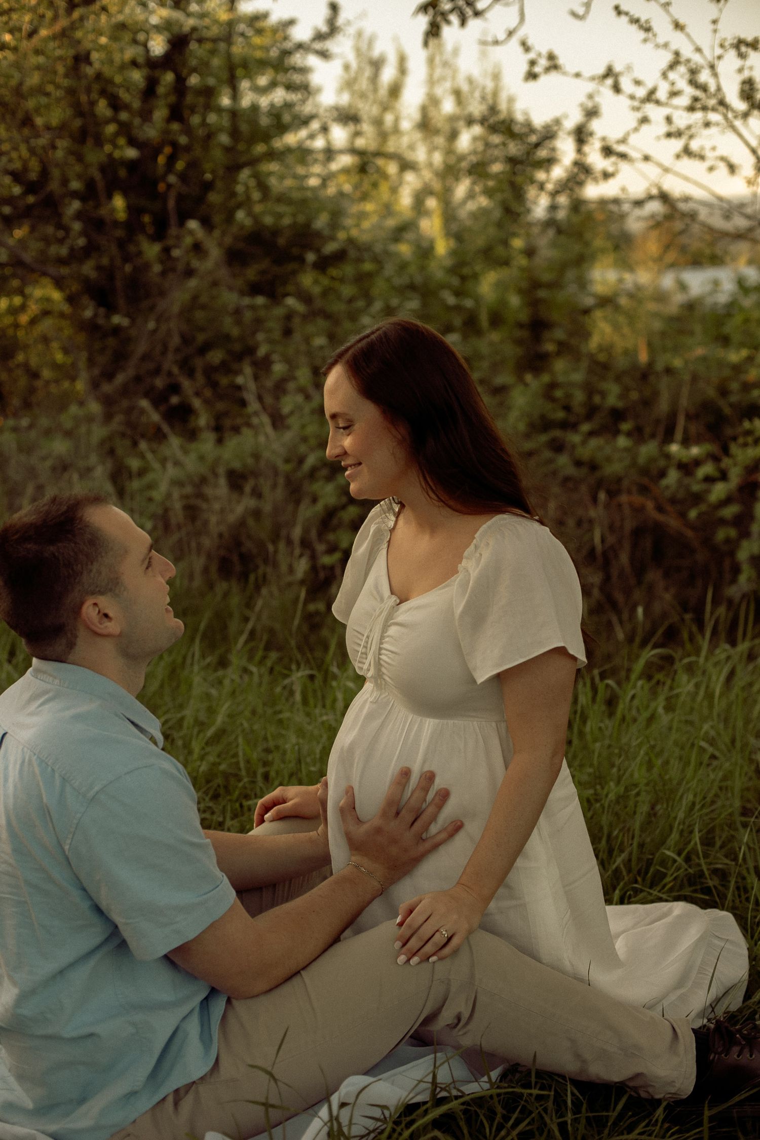 A woman leans against her partner, he holds her belly and looks up lovingly to her while they pose for photos in a cinematic, moody field