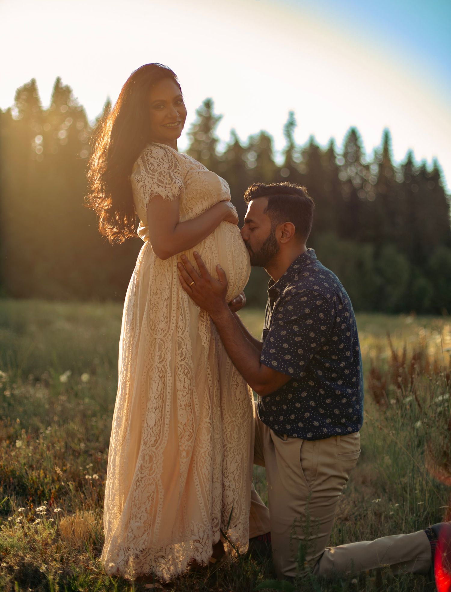 A man leans down to kiss his pregnant wife's belly in a sunny field