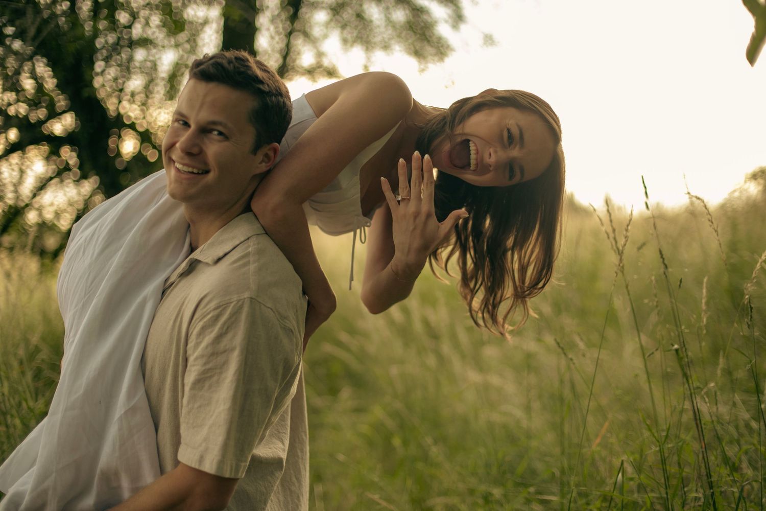 A woman is held over the shoulder of her partner while excitedly presenting her engagement ring.