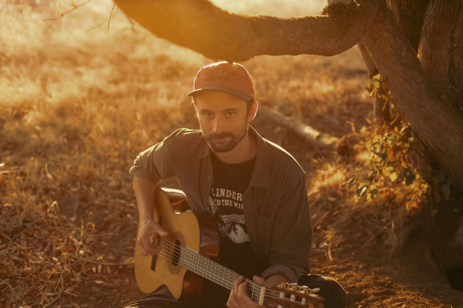 A man playing guitar under a tree at sunset in a dry grassy field.
