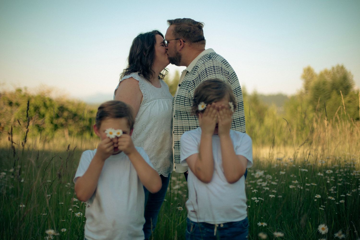 A mother and father kissing in a field of flowers while their two sons look away and hold flowers in front of their eyes.