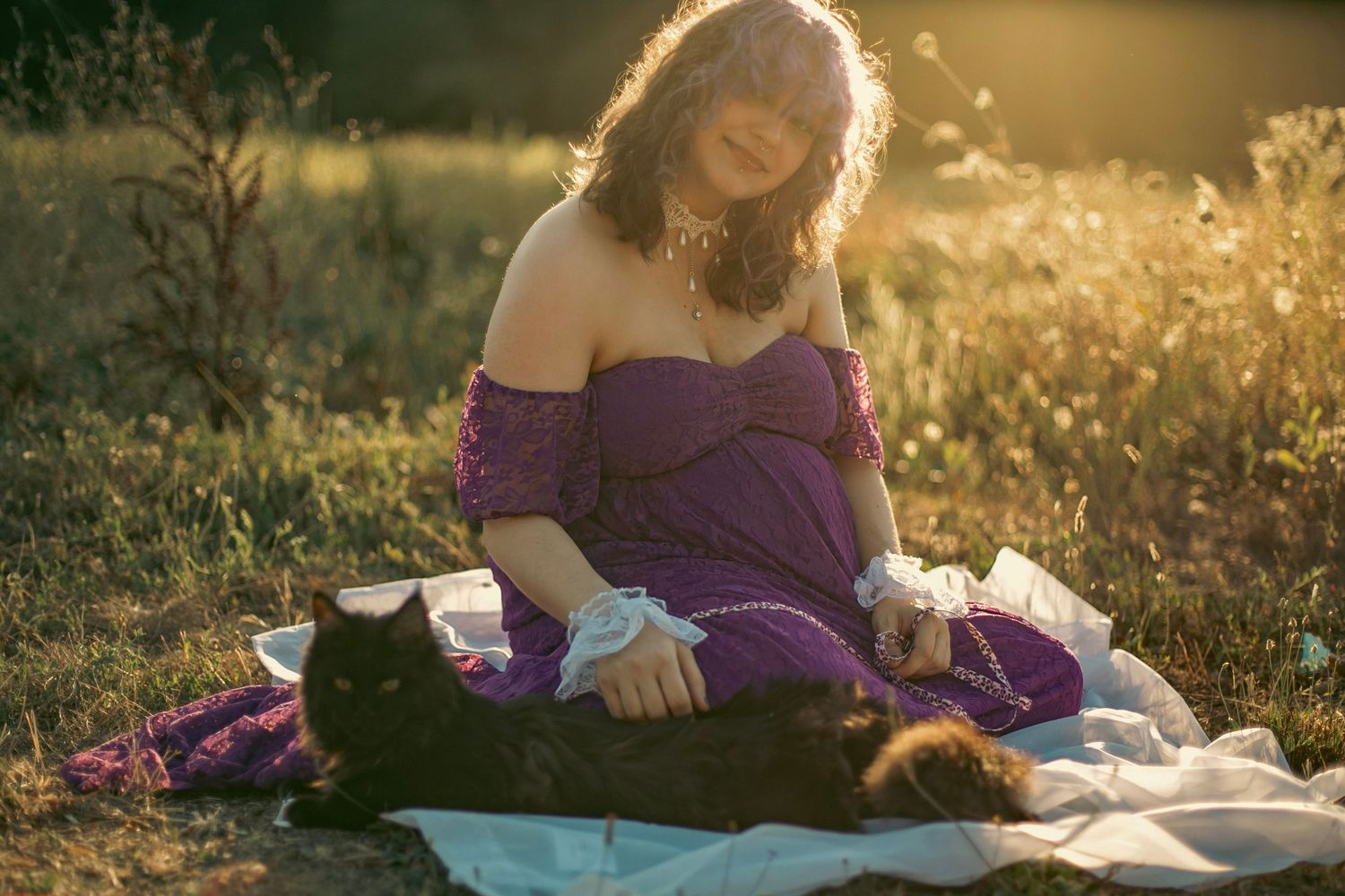 A woman in a purple dress petting a fluffy, black cat while sitting on a blanket in a sunset-lit flower field.