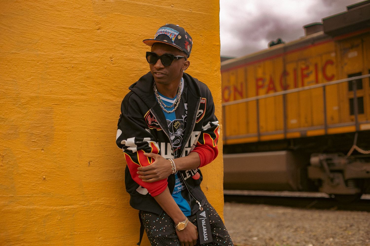 A young man wearing designer clothes poses in front of a yellow wall with a train going by in the background - urban modeling photoshoot