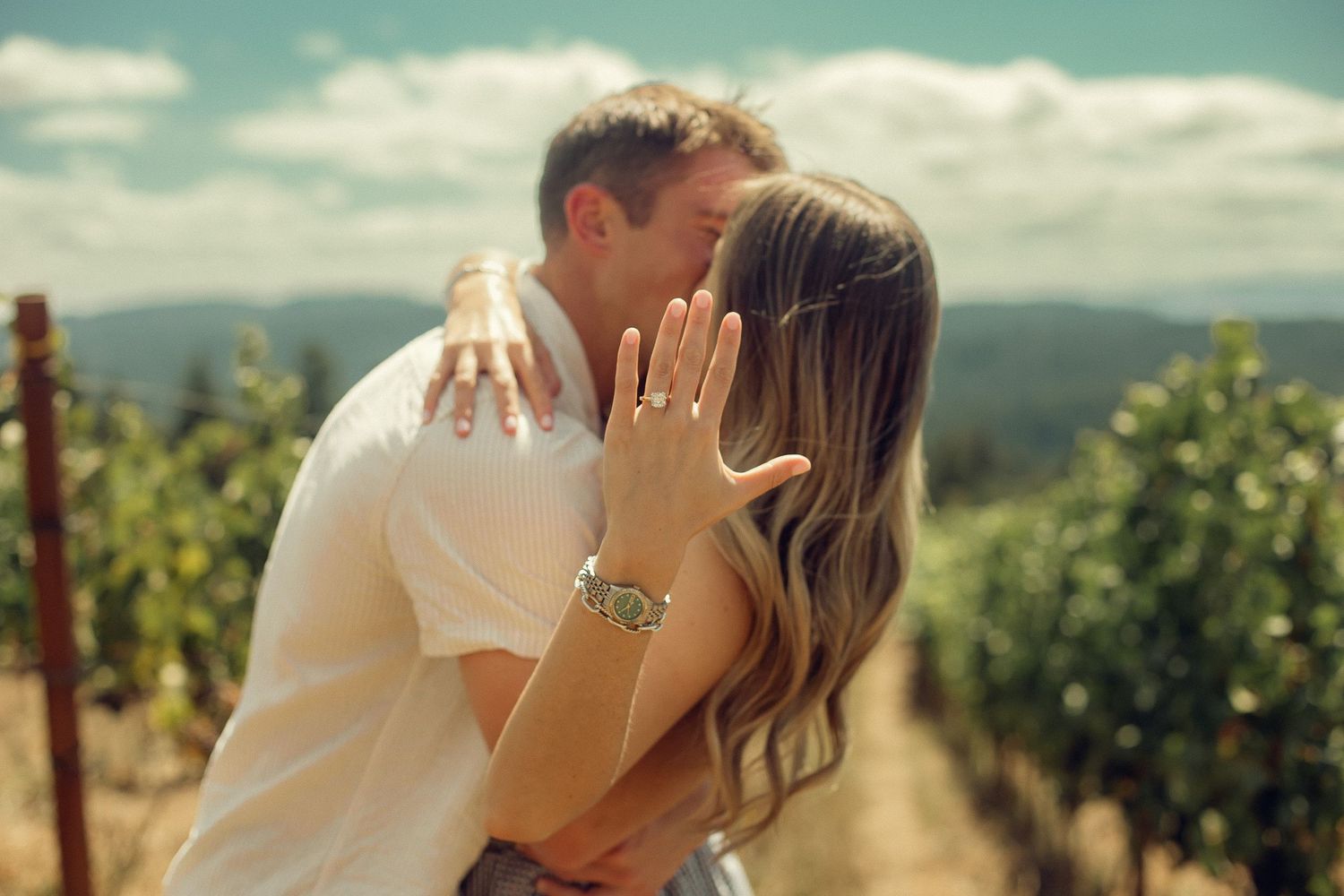 A newly engaged couple kisses and the woman holds out her hand to show off her engagement ring in blue-sky, sunny vineyard in Oregon.