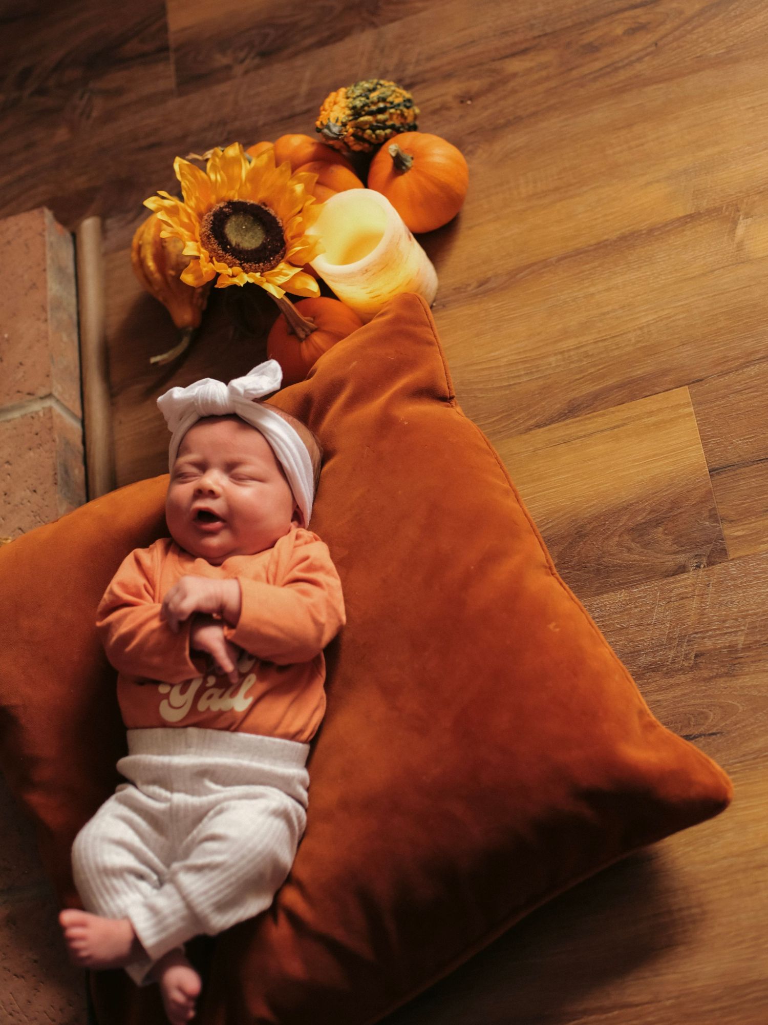 A newborn baby laying on an orange pillow with fall flowers and pumpkins behind them.