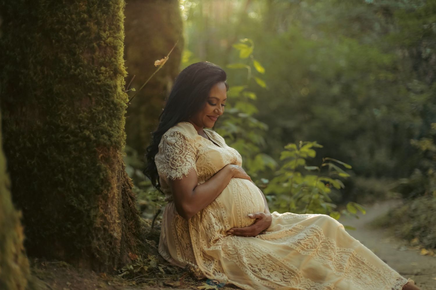 A maternity photo for a woman in a white dress holding her belly and sitting against a mossy, green tree in a lush forest.