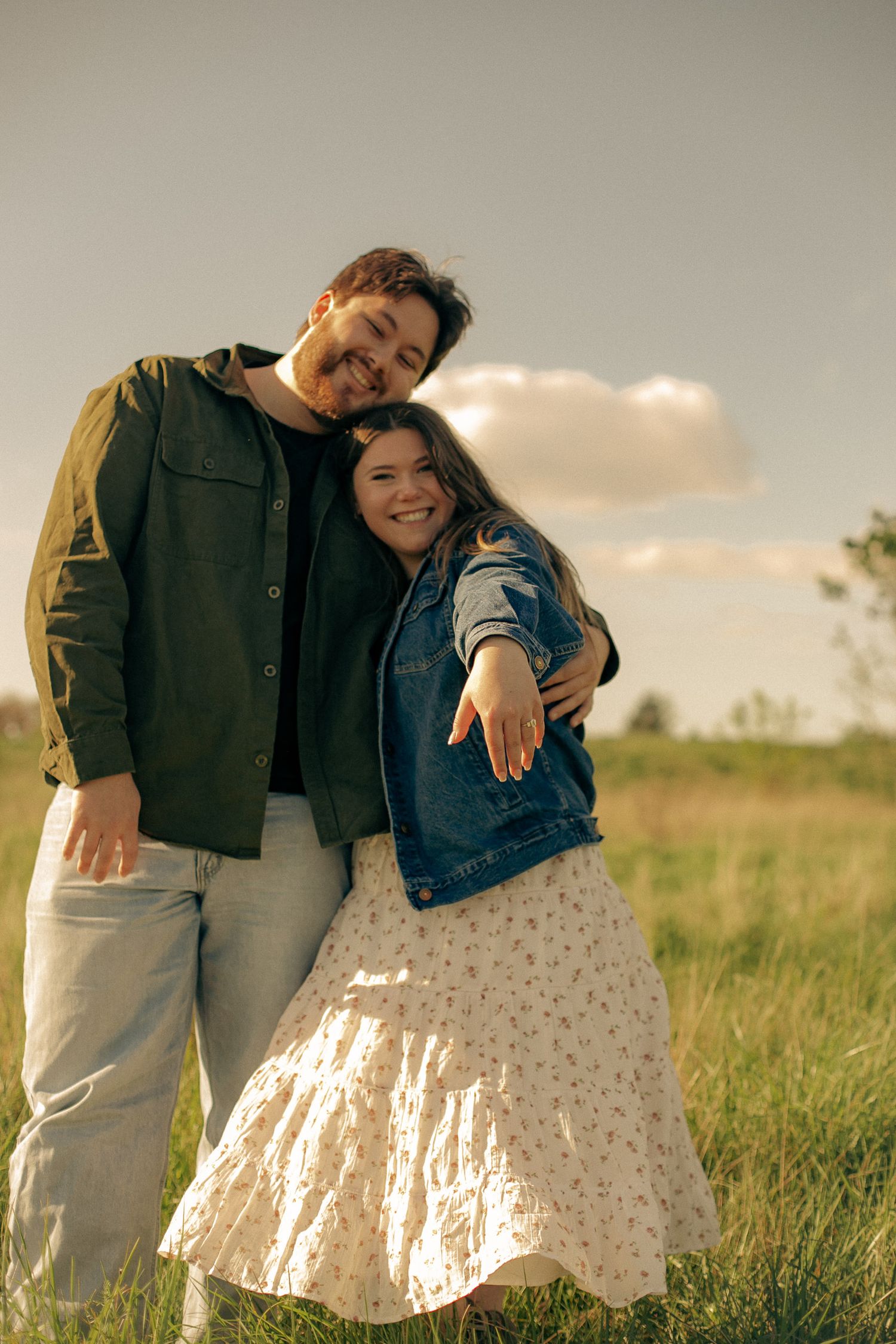 A young couple pose with the woman's hand showing her engagement ring in an open green field, after a proposal photoshoot
