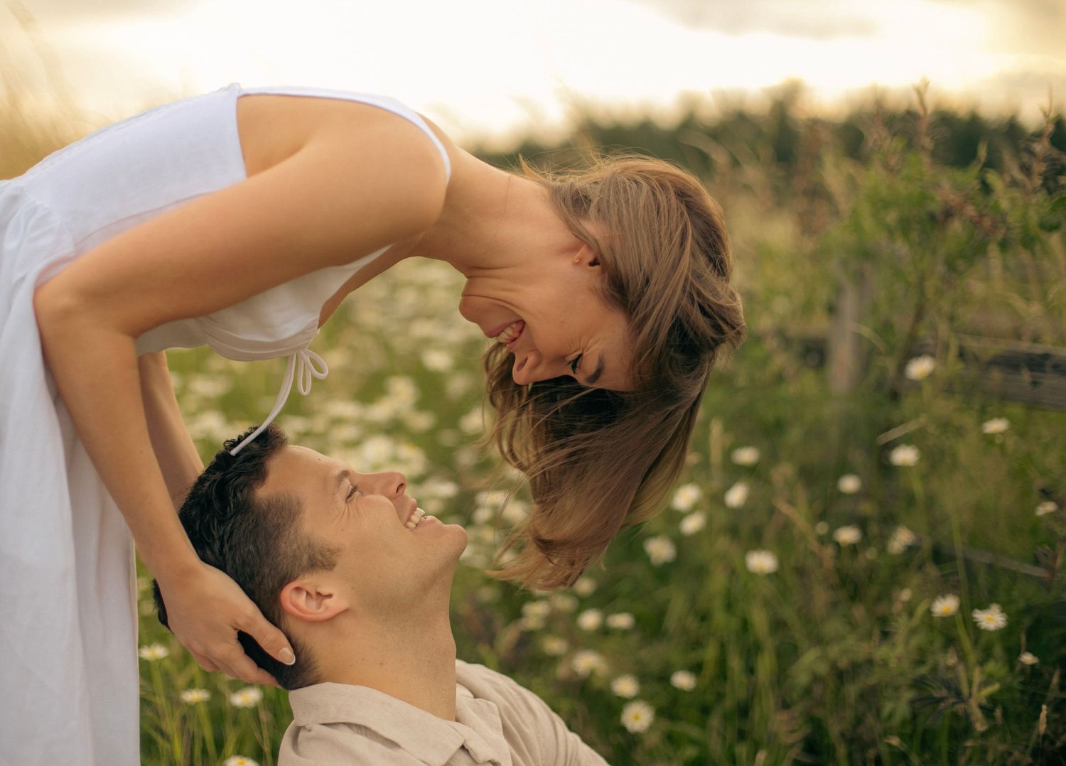A man and woman in a field of flowers, the woman is bending over the man and they are staring into each other's eyes smiling