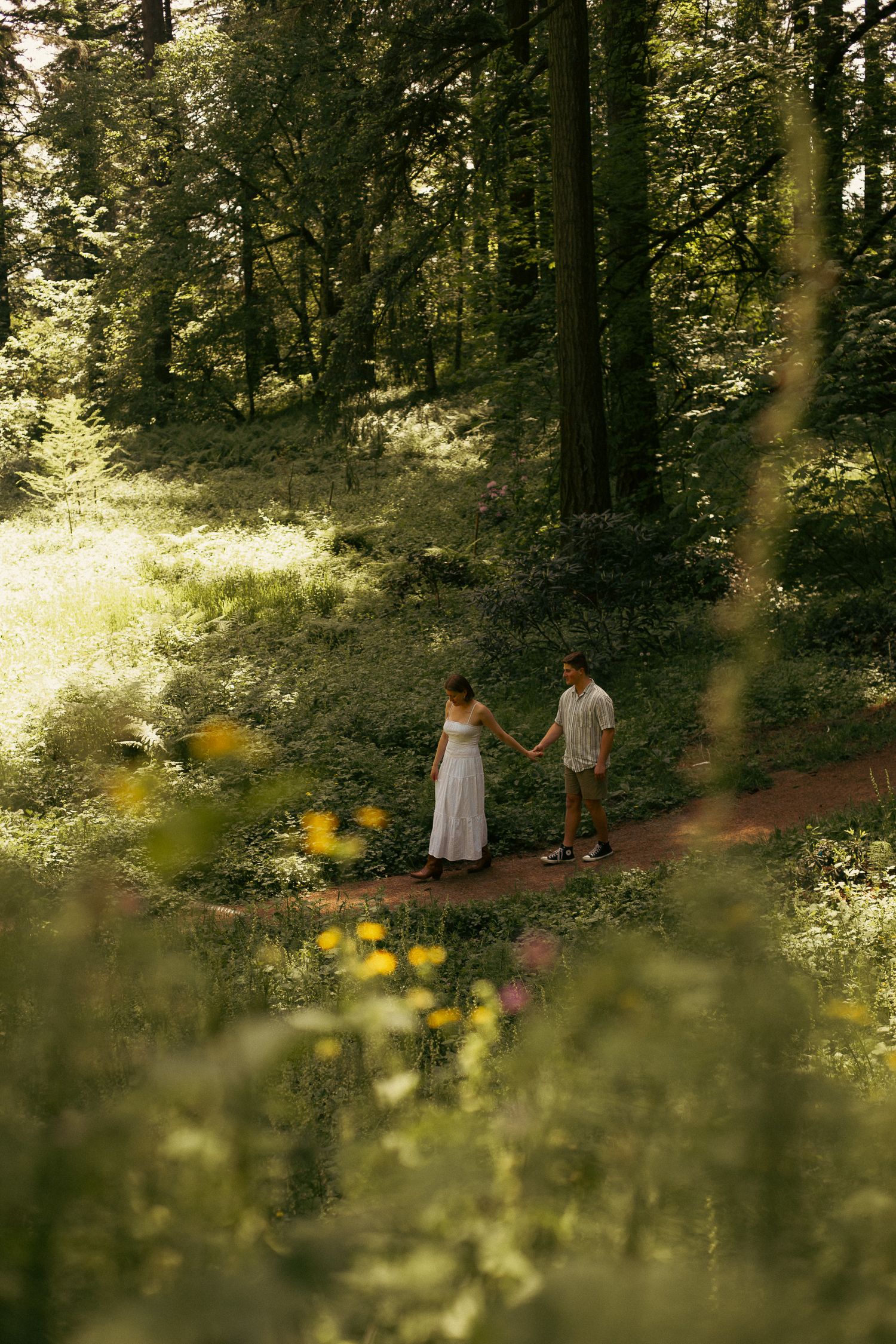A young couple walks hand-in-hand through a sun-lit forest with flowers in the foreground for an engagement photoshoot