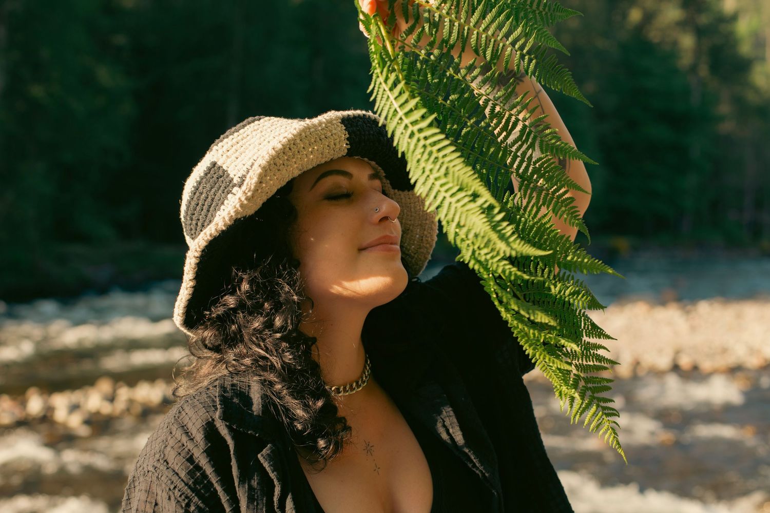 A woman with black curly hair and a hat standing in front of a river holding a fern in front of her face.