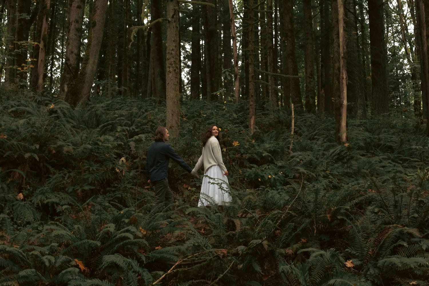 A couple holds hands while the woman is leading the man walking on a trail in a moody PNW forest